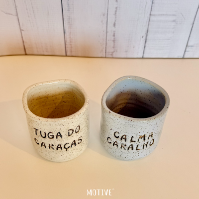 Dois copos cerâmicos com textos em português sobre superfície branca e fundo madeira