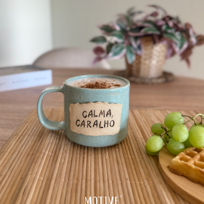 Caneca azul com frase CALMA, CARALHO sobre esteira de bambu numa mesa com uvas e waffle
