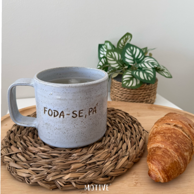 Caneca cerâmica cinza com frase gravada, suporte de palha, croissant e planta em vaso trançado