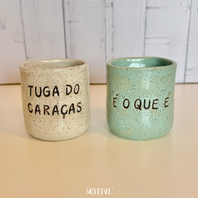 Dois copos de cerâmica com inscrições em português sobre mesa clara e fundo de madeira branca.