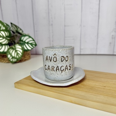 Caneca e pires em cerâmica com texto sobre tábua de madeira, planta ao fundo