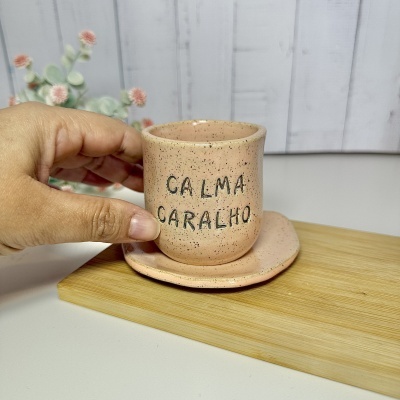 Caneca rosa em cerâmica com texto CALMA CARALHO e prato correspondente, segurada por mão.