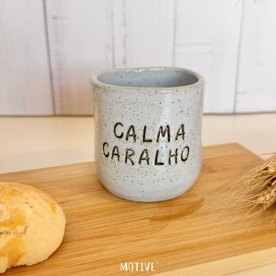 Caneca de cerâmica cinzenta com texto CALMA CARALHO sobre tábua de madeira com pão e espigas de trigo