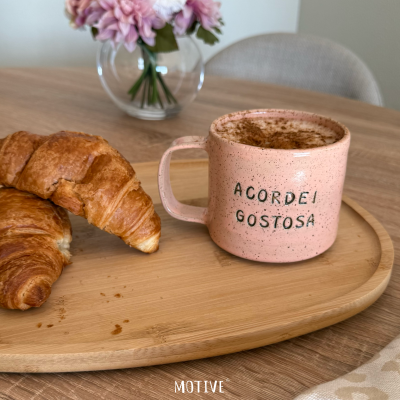 Caneca rosa com texto ACORDEI GOSTOSA e croissants numa bandeja de madeira