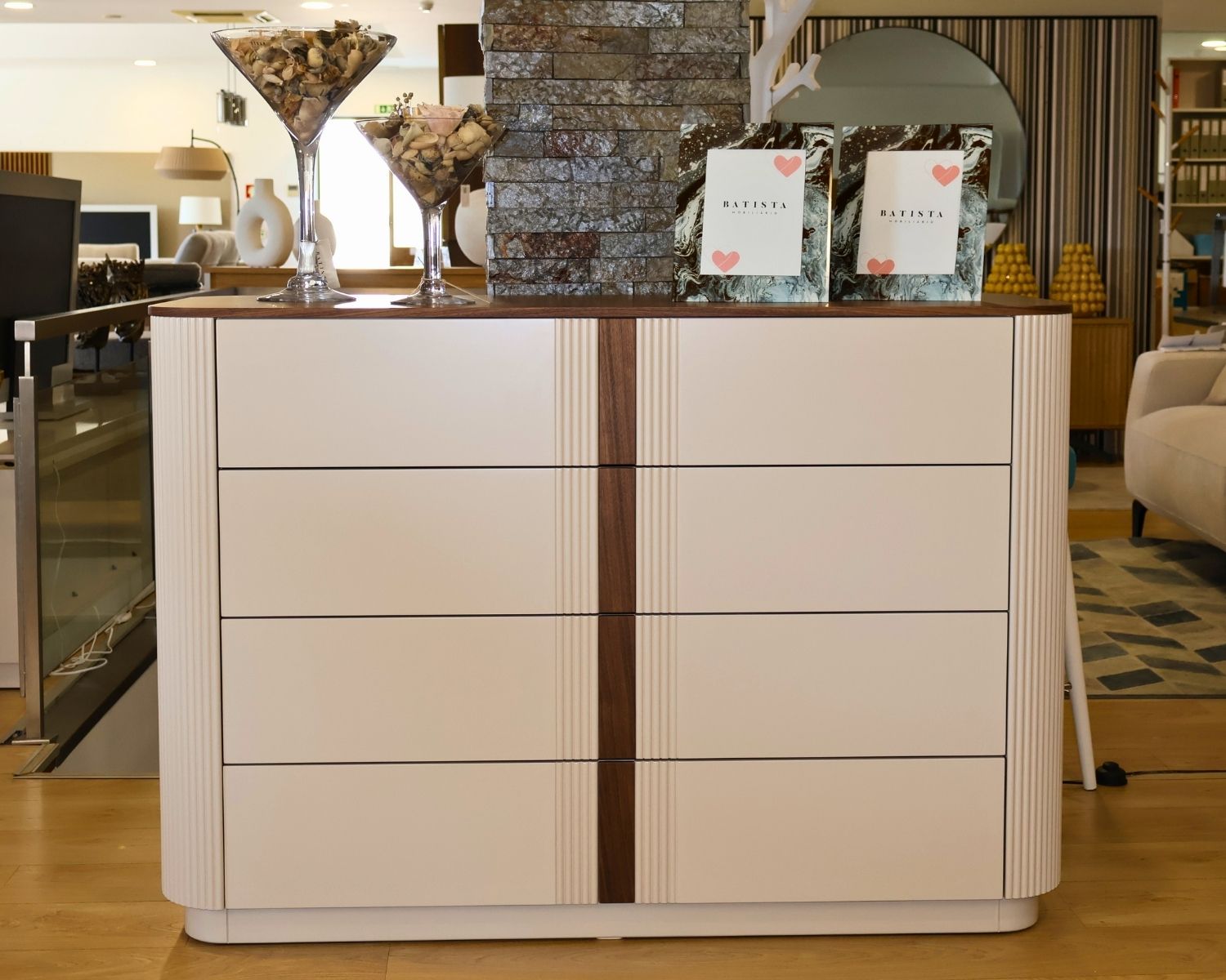 Cómoda GYO com 4 gavetas Cómoda branca com detalhes em madeira, copos decorativos cheios de conchas e quadros decorativos com texto BATISTA.
