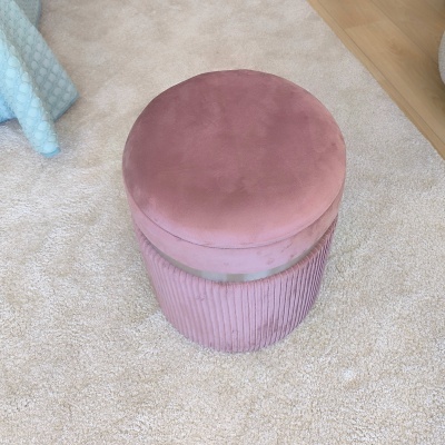 Pufe redondo rosa de veludo em carpete bege junto a chão de madeira e cama com cobertor azul