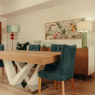 Mesa de jantar de madeira com base branca em X e cadeiras verdes, com iluminação e quadro decorativo na parede.