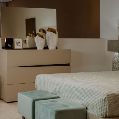 Quarto com cama bege, pufes verdes, cómoda bege, espelho e vasos decorativos