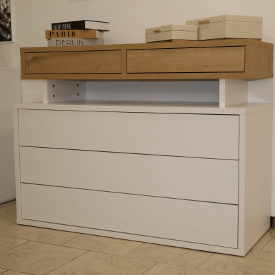 Cômoda branca com gavetas e extensão em madeira clara com caixas e livros em cima