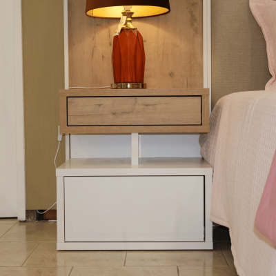 Mesa de cabeceira em madeira clara e branca com candeeiro laranja ao lado de cama com colcha branca e manta rosa.