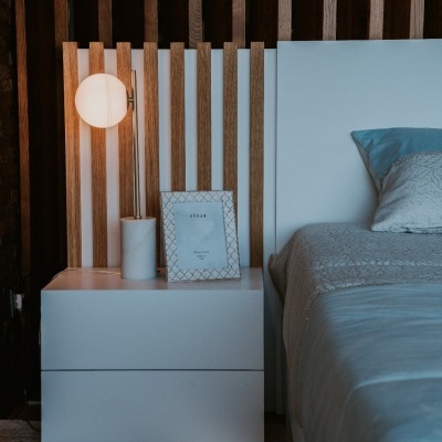 Mesa de cabeceira branca ao lado de cama com cabeceira branca e almofada azul, lâmpada e moldura de fotografia, parede de madeira ao fundo