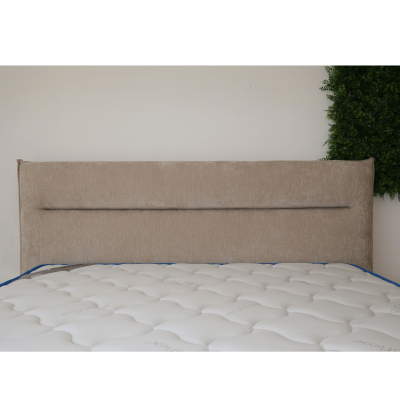 Cabeceira estofada bege com colchão branco acolchoado e detalhes azuis, parede branca e plantas verdes atrás