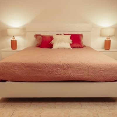 Cama branca com colcha rosa e almofadas, mesas de cabeceira com candeeiros laranja, chão de azulejo bege