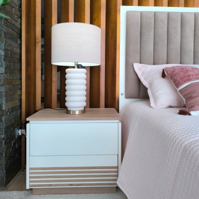 Mesa de cabeceira branca e madeira com abajur ao lado de cama com cabeceira estofada e roupa de cama rosa.