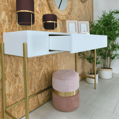 Secretária branca suspensa com pernas douradas, banco rosa redondo com faixa dourada, caixas decorativas roxas e plantas verdes num ambiente interior