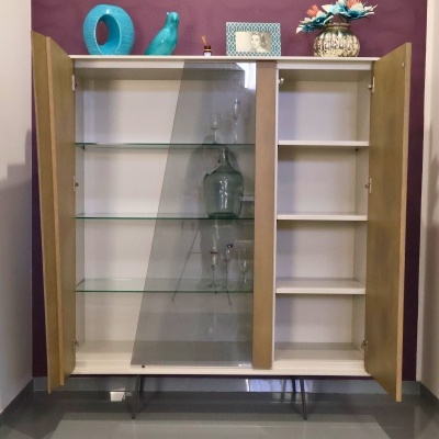 Móvel de exposição com portas madeira clara, vidro inclinado, prateleiras em vidro e madeira branca, decorado com vaso, moldura fotográfica, escultura azul e jarro de vidro.