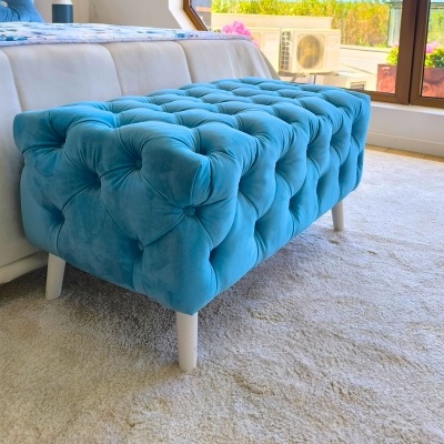 Pufe estofado azul de veludo com pernas brancas numa divisão de quarto.