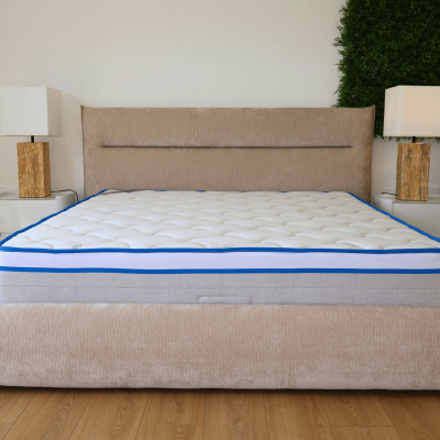 Cama com colchão branco de bordas azuis e cabeceira estofada bege numa sala com duas mesas de cabeceira e candeeiros