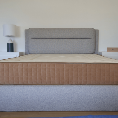 Cama com cabeceira cinzenta e colchão bege com laterais castanhas, mesas de cabeceira brancas com candeeiro e caixa