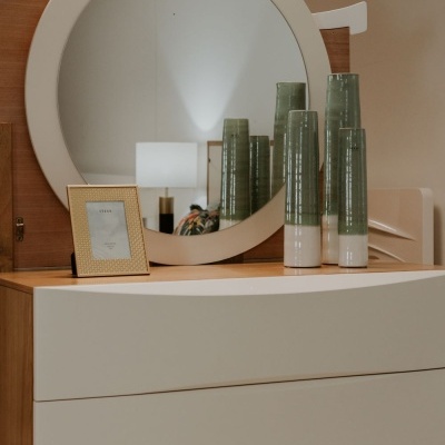 Comoda branca com espelho redondo, vasos cerâmicos verdes e porta-retrato dourado