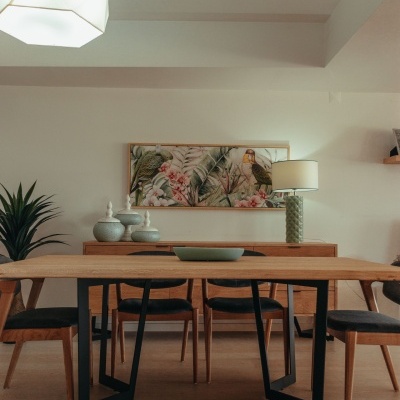 Mesa de jantar em madeira clara com cadeiras pretas num espaço interior decorado com quadro tropical e candeeiro