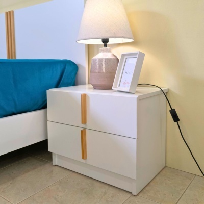 Mesa de cabeceira branca com candeeiro e moldura, próxima de cama com roupa azul