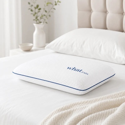 Travesseiro branco com borda azul e texto what.CASA sobre cama branca com almofada e vaso decorativo