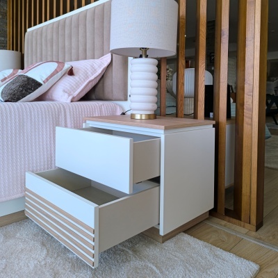 Mesa de cabeceira branca com gavetas abertas e candeeiro ao lado de cama com cabeceira bege.