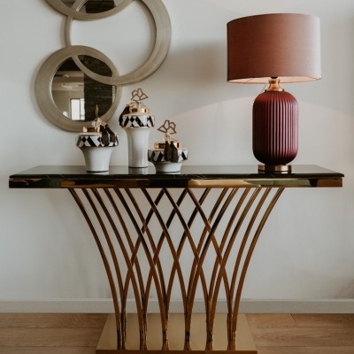 Consola moderna com tampo preto e base dourada, esculturas em branco e preto e lâmpada vermelha com candeeiro rosa, espelhos redondos na parede branca.