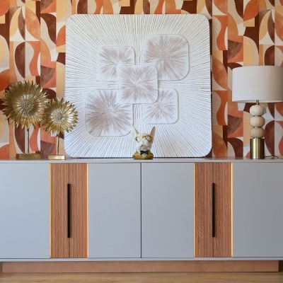 Aparador cinza claro com decoração dourada e lâmpadas, em ambiente com papel de parede geométrico laranja.