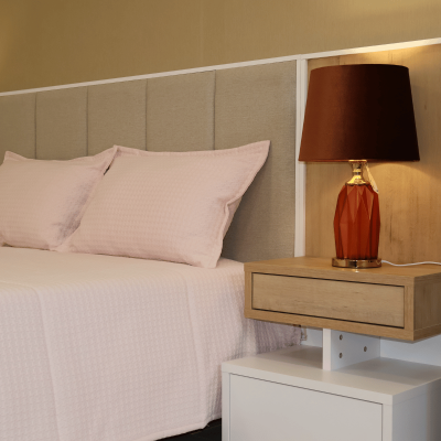 Cama com cabeceira cinza clara, roupa de cama rosa, criado-mudo com gaveta e candeeiro vermelho