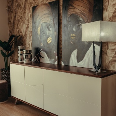 Móvel de armazenamento creme e madeira com pinturas de mulheres com turbantes e lâmpada em ambiente com papel de parede de folhas