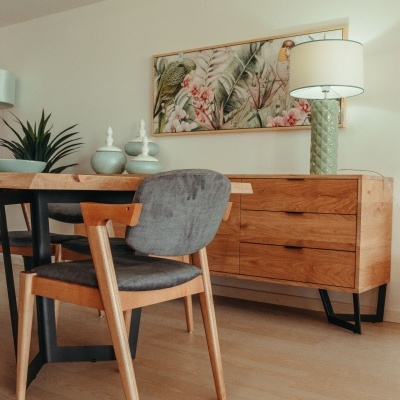 Cadeira de madeira com estofos cinzentos junto a mesa redonda e aparador de madeira com candeeiro e quadro decorativo