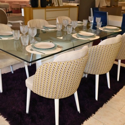 Mesa de jantar de vidro com cadeiras estofadas bege e padrão geométrico amarelo, sobre tapete roxo