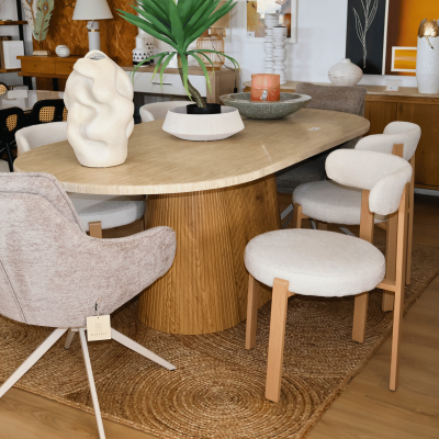 Mesa de centro oval em madeira clara com cadeiras estofadas claras e elementos decorativos no tampo.