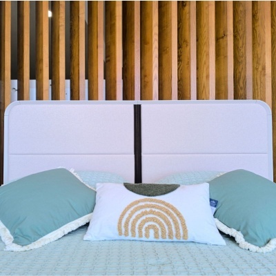Cabeceira de cama cinza claro com almofadas decorativas verdes e branca com padrão de arco-íris, colcha azul claro e parede de ripas de madeira ao fundo.