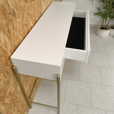 Secretária branca com gaveta aberta e pernas douradas perto de parede de madeira e plantas em vasos