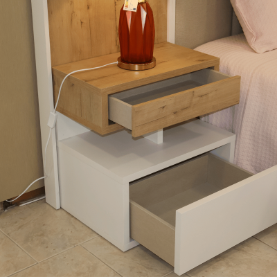 Mesa de cabeceira com duas gavetas, uma branca e outra de madeira clara, junto a cama com colcha cor de rosa e candeeiro vermelho.