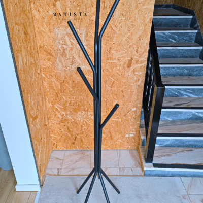 Cabide de pé preto fosco de design minimalista em forma de árvore junto a parede de aglomerado com texto BATISTA MOBILIÁRIO e escadas de mármore cinza.