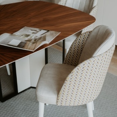 Cadeira estofada cinza claro com padrão geométrico atrás, ao lado de mesa de madeira escura com revista