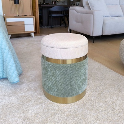 Pufe cilíndrico verde com topo creme e faixa dourada, colocado sobre tapete claro em sala com sofá e móvel