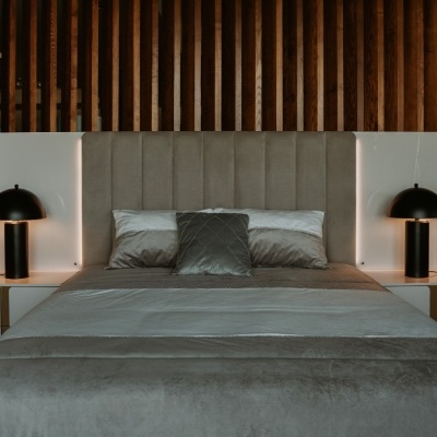 Cama de cabeceira alta cinza com candeeiros pretos em mesas brancas, decorada com almofadas e parede de madeira atrás.