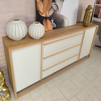 Aparador moderno em madeira clara com gavetas e portas brancas, vasos brancos e objeto dourado em cima