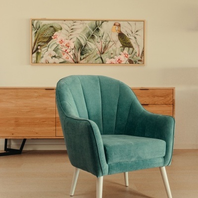 Cadeira verde água acolchoada com pernas brancas diante de móvel de madeira e quadro com papagaios