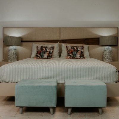Quarto com cama grande, cabeceira estofada bege, roupa de cama cinza e almofadas coloridas com duas mesas de cabeceira e puffs azuis claros.