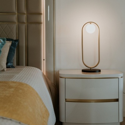 Mesa de cabeceira branca com candeeiro dourado ao lado de cama com cabeceira castanha e colcha branca