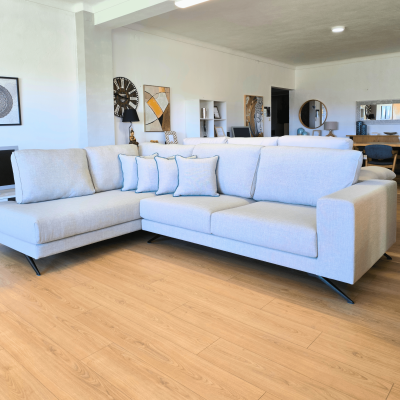 Sofá de canto cinza claro com almofadas e pernas metálicas pretas numa sala moderna com chão de madeira.