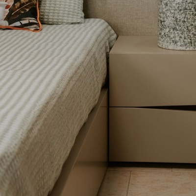 Mesa de cabeceira bege fosco ao lado de cama com colcha cinza e almofada decorativa