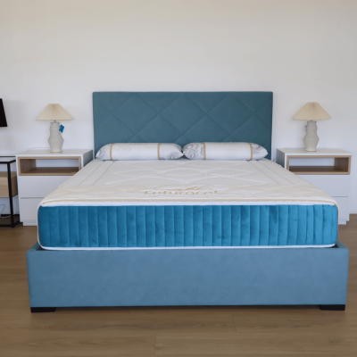 Cama estofada azul com colchão branco e duas almofadas cilíndricas, mesas de cabeceira com lâmpadas em quarto minimalista