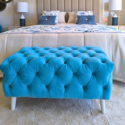 Poltrona estofada azul com botão capitonê e pernas brancas num quarto ao lado de cama